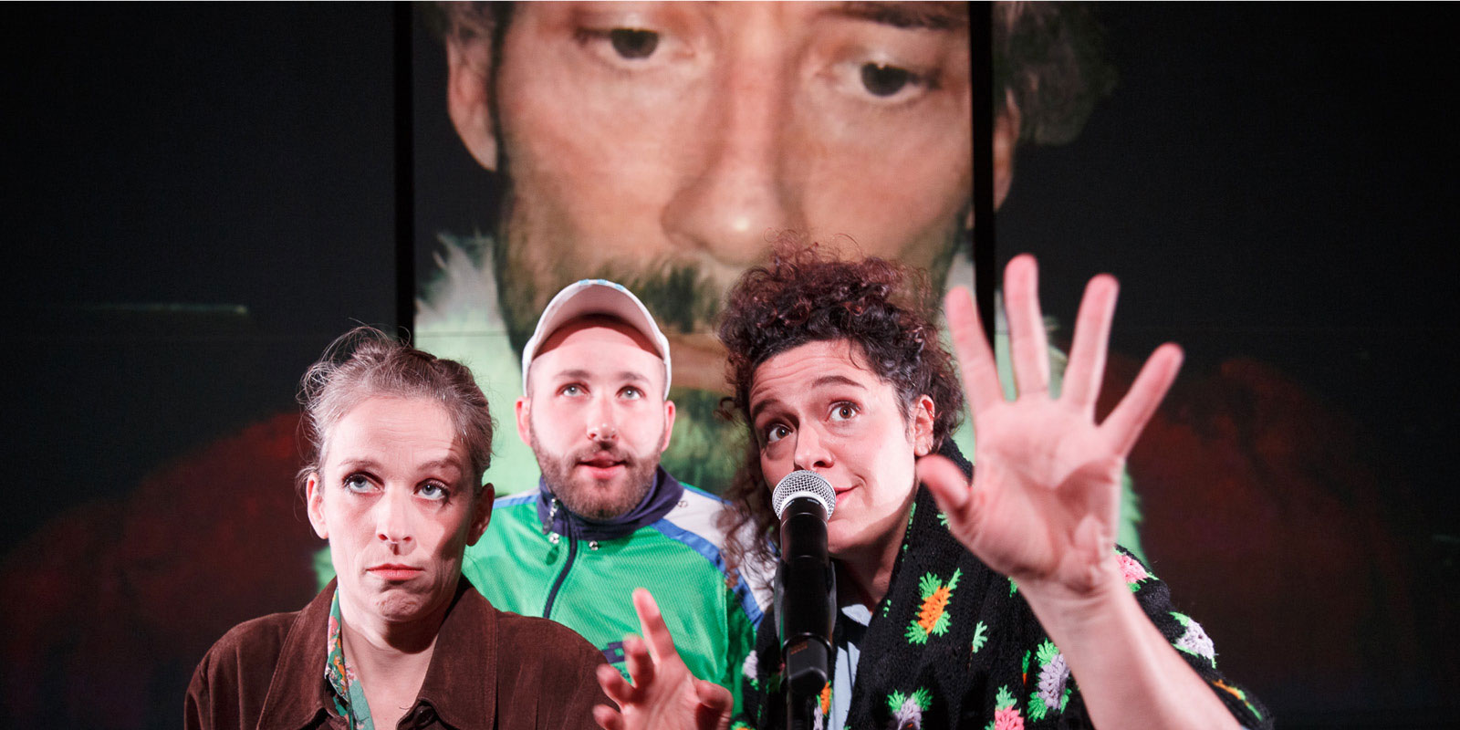 photo issue du spectacle "Grand Pays" montrant trois comédiens au premier plan derrière un micro. Un visage est projeté en grand en arrière plan.