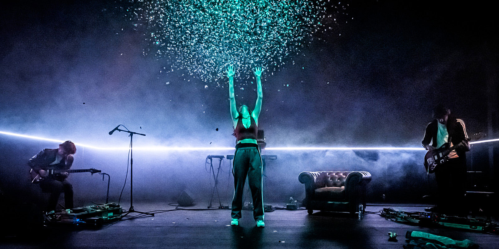 Photo issue du spectacle "Iphigénie à Splott" avec une comédienne au centre lançant des confettis en l'air, entourée de deux guitaristes de chaque côté.
