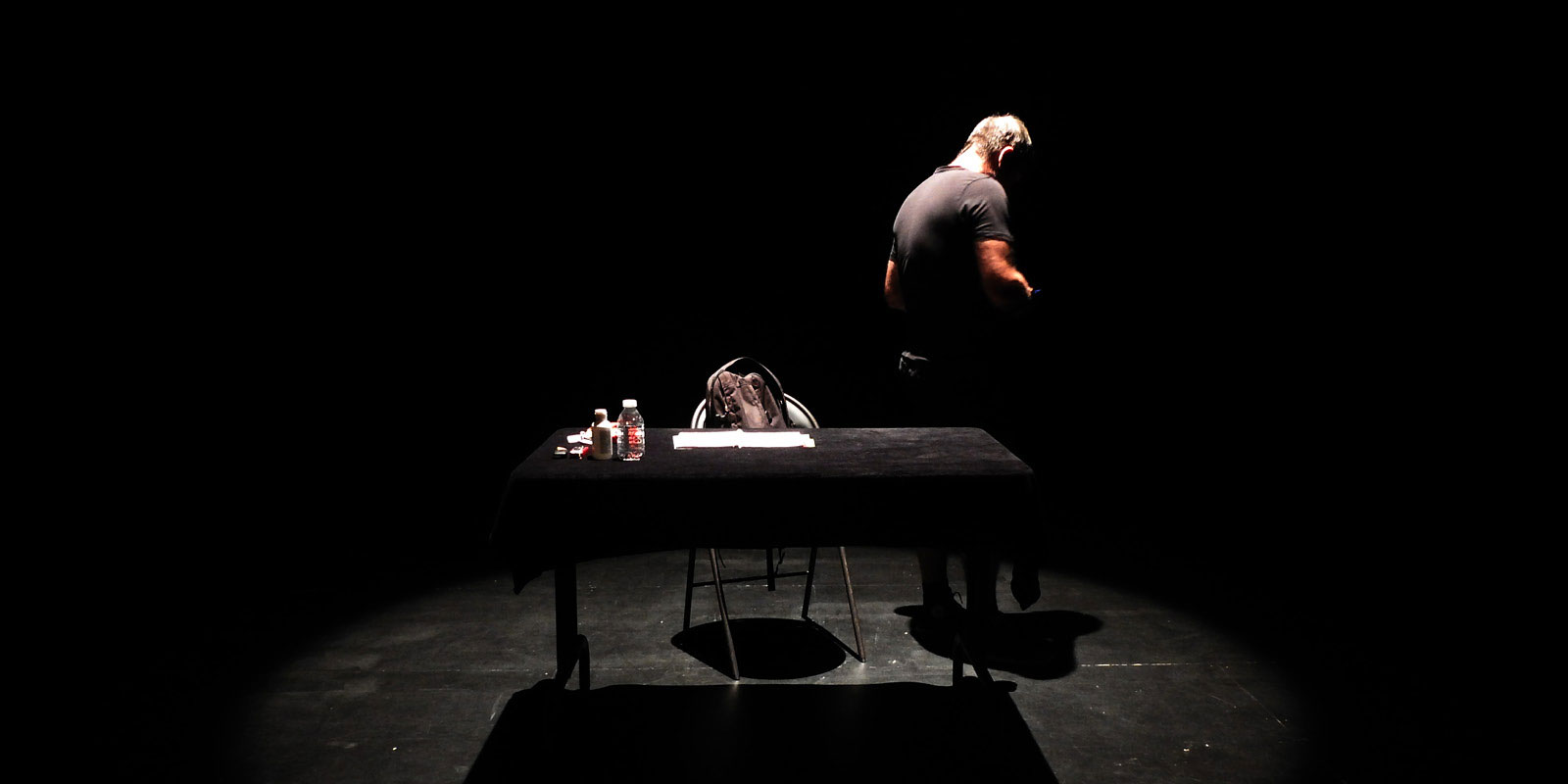 Photo d'un homme, habillé en noir, de dos derrière une table, issue du spectacle "Rencontre avec Michel B.